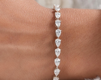 May include: A silver-toned tennis bracelet featuring a continuous row of clear, pear-shaped stones set in a metallic frame. The bracelet is designed with a flexible link structure, showcasing a series of identical teardrop-cut gems arranged vertically along the wrist.