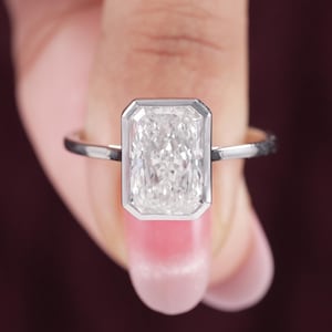 May include: A silver-toned ring featuring a large, rectangular, radiant-cut clear gemstone set in a smooth, polished bezel setting. The band is thin and metallic, providing a clean, reflective surface that highlights the central stone's facets.