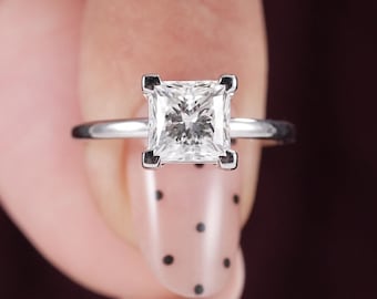 May include: A silver-coloured ring featuring a single square-cut princess diamond held in a four-prong setting. The band is smooth and polished, showcasing a clean and simple design that highlights the faceted gemstone at the centre of the piece.