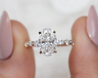 May include: A ring featuring a large oval-cut center stone held by four prongs, set on a thin band adorned with smaller round and marquise-cut stones along the sides. The metal band appears to have a warm, metallic tone, reflecting light.