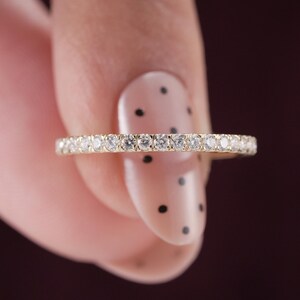 May include: A thin gold band ring featuring a continuous row of small, round, clear faceted stones set along the entire outer circumference. The metal has a polished yellow gold finish, and the stones are arranged in a uniform, prong-set design.