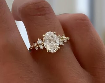 Può includere: Un anello in oro con una grande pietra centrale a taglio ovale, affiancata da un gruppo di tre pietre più piccole a taglio rotondo su ciascun lato. La fascia è sottile e lucida, con le pietre fissate in una montatura a griffe.
