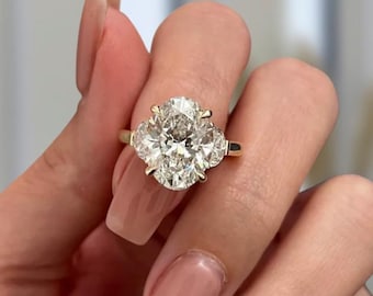 May include: A gold ring featuring a large oval-cut center stone flanked by two smaller, semi-circular stones on each side. The setting is crafted from polished yellow gold, holding the clear, faceted stones securely in place with multiple prongs.