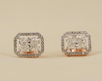 May include: A pair of stud earrings featuring a central rectangular radiant-cut stone surrounded by a halo of small round pavé-set stones. The earrings are set in a metallic frame with a warm-toned base visible beneath the central stones.