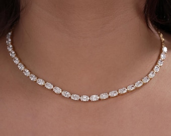 May include: A gold-toned tennis necklace featuring a continuous row of sparkling, oval-cut clear stones. The necklace sits flush against the neck, showcasing a uniform arrangement of faceted gems set in a delicate metal chain with a polished finish.