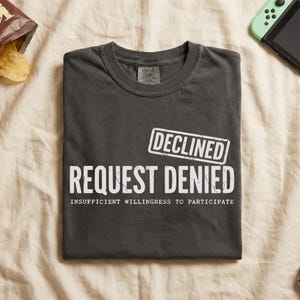 Op de afbeelding: Donkergrijs t-shirt met de witte tekst "REQUEST DENIED" en "DECLINED", evenals de zin "INSUFFICIENT WILLINGNESS TO PARTICIPATE". Een zak chips en een spelconsole zijn op de achtergrond.