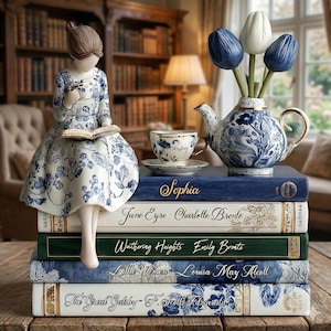 May include: A decorative scene featuring a porcelain figurine reading on a stack of books. The books are titled "Sophia", "Jane Eyre", "Wuthering Heights", "Little Women", and "The Great Gatsby". A teapot vase with tulips and a teacup are also present.