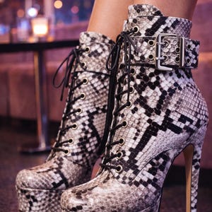 Könnte beinhalten: Ankle-Boots mit Plateau und hohem Absatz in Schlangenmuster in Schwarz, Weiß und Grau. Die Stiefel haben eine Schnürung, einen Schnallenverschluss und einen Stiletto-Absatz.