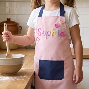 Op de afbeelding: Een roze schort met een marineblauwe zak en bandjes. De naam "Sophia" is met kleurrijk garen op de voorkant geborduurd. Een houten lepel wordt vastgehouden in de buurt van een mengkom, wat wijst op een bakthema.