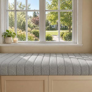 May include: A light gray upholstered bench with a vertical channel-tufted design and a textured fabric finish. The bench features a solid light-colored wooden base with two rectangular cabinet panels positioned beneath a large window.