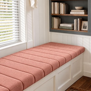May include: A long, rectangular window bench featuring a textured, dusty rose-colored upholstered cushion with horizontal channel tufting. The base is constructed from white wood with a paneled design, positioned beneath a window with white blinds.