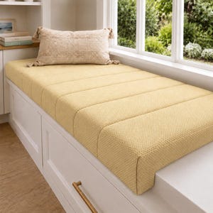 May include: A rectangular, textured, light yellow bench cushion with vertical channel tufting. A matching light beige, rectangular throw pillow with decorative tassels at each corner rests on top of the cushion, which is placed on a white base.