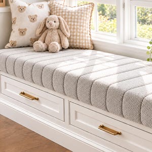 May include: A white window bench with two drawers featuring gold-toned metal handles. The bench is topped with a thick, light gray, vertically tufted cushion. A cream-colored bear-patterned pillow and a beige gingham pillow sit on the bench.