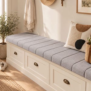 May include: A white wooden storage bench featuring three drawers with dark metal cup pulls and a thick, light gray tufted cushion on top. The bench is rectangular with a clean frame design and provides seating space with integrated storage.