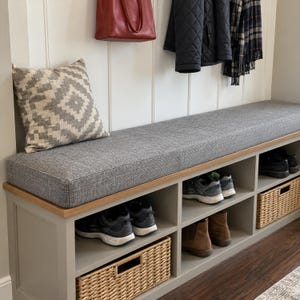 May include: A gray entryway bench with a wooden top, a thick gray fabric cushion, and a square throw pillow with a gray and white geometric pattern. The base features open cubbies containing various pairs of shoes and two woven wicker baskets.