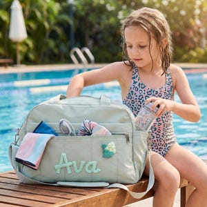 Könnte beinhalten: Eine hellgrüne Reisetasche mit dem Namen "Ava" auf der Vorderseite gestickt, neben einer floralen Applikation. Die Tasche ist offen und zeigt Handtücher und Accessoires. Ein Kind in einem Badeanzug mit Leopardenmuster hält eine Wasserflasche.