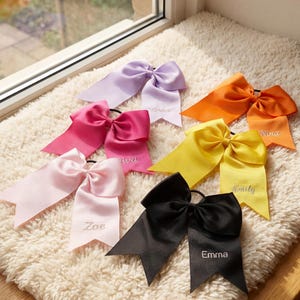 Peut inclure: Plusieurs nœuds pour cheveux en satin de couleurs vives : rose, violet, jaune, orange et noir. Chaque nœud porte un prénom brodé sur le ruban. Les nœuds sont disposés sur une surface blanche et moelleuse, près d'une fenêtre.