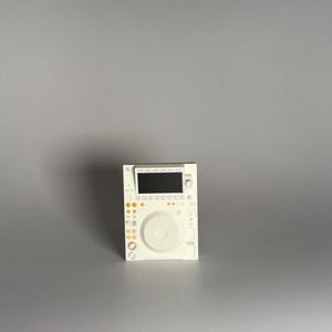 Pode incluir: Um controlador de DJ branco com tela preta e botões laranja. O controlador possui um jog wheel circular e vários botões e botões. O dispositivo foi projetado para mixar e reproduzir música.