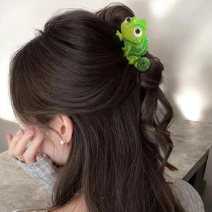 Peut inclure: Une pince à cheveux en forme de caméléon vert vif avec une queue enroulée et de grands yeux blancs. Le caméléon est placé sur des cheveux châtain foncé, maintenant une mèche de cheveux dans une coiffure demi-attachée grâce à sa pince intégrée.