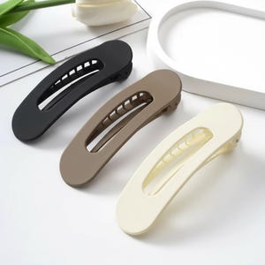 Könnte beinhalten: Drei große Haarspangen in Schwarz, Taupe und Creme. Jede Spange hat eine gebogene, längliche Form mit einem mittigen Ausschnitt und einer Reihe von Zähnen an einer Seite. Die Spangen bestehen aus einem matten Material und dienen zur Befestigung der Haare.