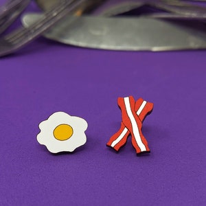 May include: A pair of wooden earrings, one shaped like a fried egg with a yellow yolk and the other shaped like two pieces of bacon.