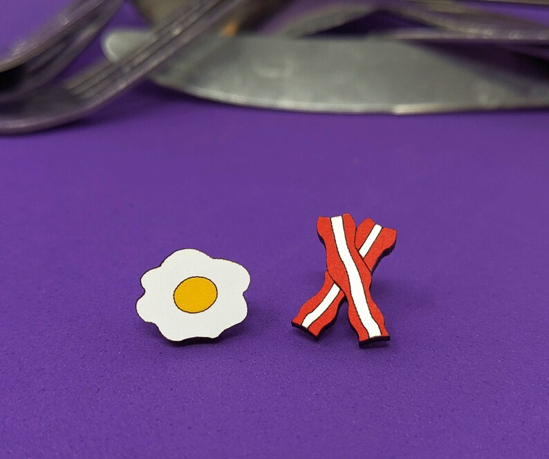 May include: A pair of wooden earrings, one shaped like a fried egg with a yellow yolk and the other shaped like two pieces of bacon.