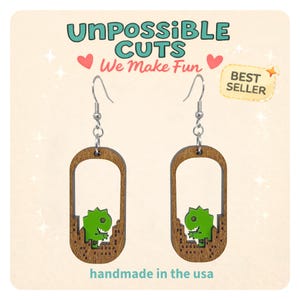 May include: Wooden earrings with a dinosaur design. The earrings are brown with a green dinosaur and the text "UnPossible Cuts We Make Fun" and "handmade in the usa". The earrings are a best seller.