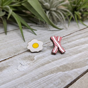 May include: Two wooden pins shaped like breakfast food. One pin is a white fried egg with a yellow yolk. The other pin is a red and white bacon strip.