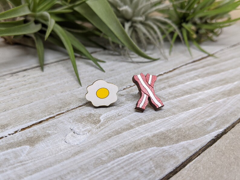 May include: Two wooden pins shaped like breakfast food. One pin is a white fried egg with a yellow yolk. The other pin is a red and white bacon strip.
