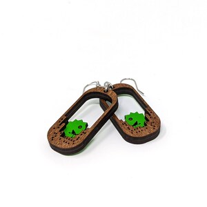 May include: A pair of wooden dangle earrings. Each earring features a dark brown, rectangular frame with a green dinosaur-shaped cutout. The earrings have silver-colored ear wires and are set against a white background.