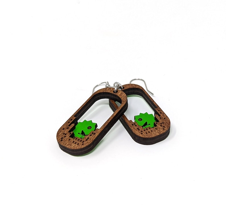 May include: A pair of wooden dangle earrings. Each earring features a dark brown, rectangular frame with a green dinosaur-shaped cutout. The earrings have silver-colored ear wires and are set against a white background.