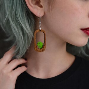 May include: Wooden dangle earrings with a rectangular frame and a green dinosaur design. The earrings have a silver hook and a detailed wood grain pattern. The dinosaur is a bright green color.