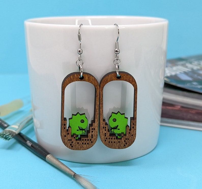 May include: Wooden dangle earrings with a retro video game dinosaur design. The earrings feature a light brown wooden frame with a green dinosaur character inside. The earrings hang from silver-colored hooks.