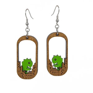 May include: A pair of wooden dangle earrings. Each earring features a brown, rectangular frame with a green dinosaur design. The dinosaur is set against a cityscape. The earrings have silver-colored fish hook ear wires.