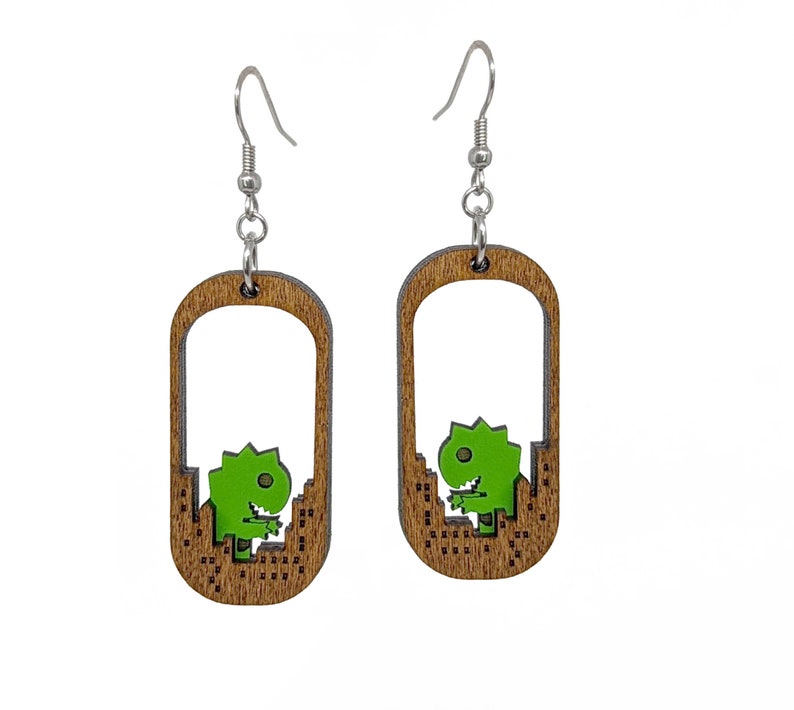 May include: A pair of wooden dangle earrings. Each earring features a brown, rectangular frame with a green dinosaur design. The dinosaur is set against a cityscape. The earrings have silver-colored fish hook ear wires.