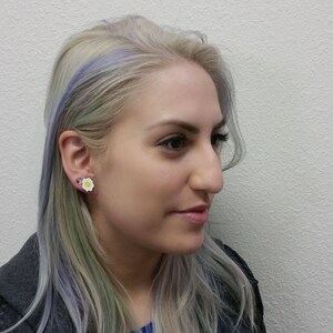 May include: A woman with light purple and green hair is wearing a white flower-shaped earring with a yellow center.
