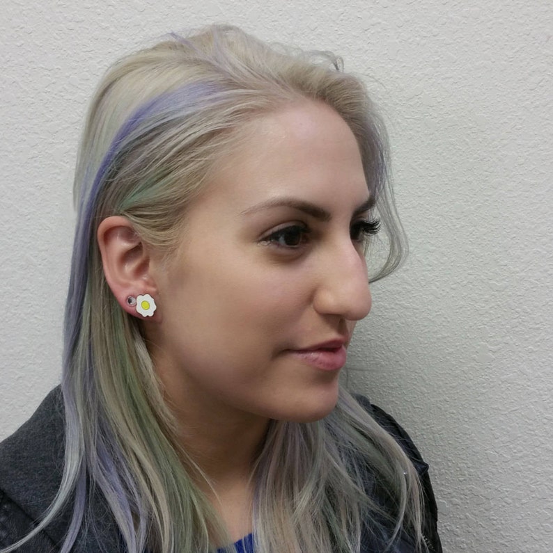 May include: A woman with light purple and green hair is wearing a white flower-shaped earring with a yellow center.