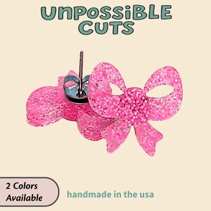 May include: Pink glitter bow earrings. The earrings are in the shape of a bow with a post backing. The text "UnPossible Cuts" is at the top of the image. The text "2 Colors Available" and "handmade in the usa" are at the bottom.