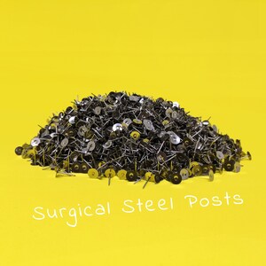 May include: A large pile of silver surgical steel earring posts on a yellow background. The text "Surgical Steel Posts" is written in white below the pile of posts.