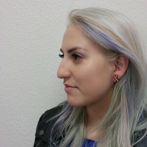 May include: A woman with light blonde hair with streaks of purple and blue, wearing a black leather jacket, is looking to the side. She is wearing red and white earrings.