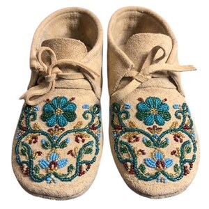 May include: A pair of beige suede moccasins with teal, blue, and red floral beadwork. The beaded design decorates the toe area, and the shoes have laces. These handcrafted moccasins are a unique footwear item.