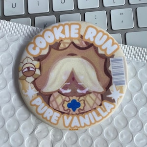 Op de afbeelding: Een ronde pin met de tekst "COOKIE RUN PURE VANILLA" in lichtbruin. De pin toont een stripfiguur met een witte baard en een blauw embleem.