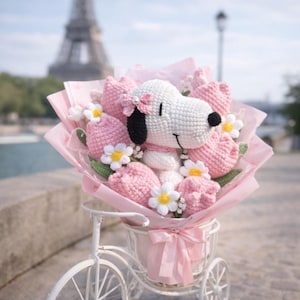 May include: A crochet bouquet with a white Snoopy plushie, pink tulips, and white daisies. The bouquet is wrapped in pink paper and presented in a white bicycle basket with a pink ribbon. The Eiffel Tower is in the background.