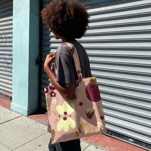 Puede incluir: Un bolso tote de color rosa claro con un patrón repetido de flores amarillas, formas marrones y acentos granate. El bolso se lleva al hombro y tiene correas a juego. El fondo es una persiana metálica gris.