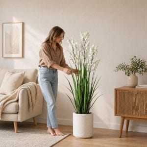 May include: A tall, artificial plant with white flowers and long green leaves in a speckled, cylindrical pot. The plant is positioned next to a person in a light-colored room with a sofa, rug, and wooden cabinet.