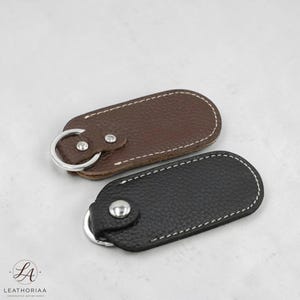 Puede incluir: Se muestran dos llaveros de cuero, uno marrón y otro negro. Cada llavero tiene un anillo de metal plateado y un cierre de botón a presión. Los bordes están cosidos con hilo blanco. El cuero tiene un aspecto texturizado.