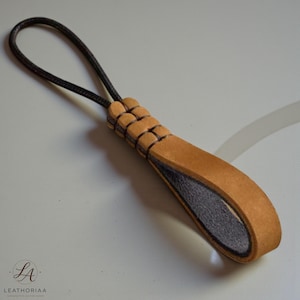 Puede incluir: Tirador de cremallera de cuero con un lazo de cordón marrón. El tirador es de cuero marrón con un interior gris oscuro y un diseño tejido en la parte superior. El logotipo "LEATHORIAA" está en la esquina inferior izquierda.