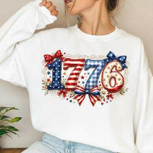 May include: White sweatshirt featuring the number "1776" in a patriotic design. The numbers are adorned with red, white, and blue patterns, bows, and floral details. A celebratory garment.