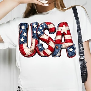 May include: White t-shirt featuring the word "USA" in a patriotic design. The letters are crafted from a patchwork of red, white, and blue fabrics, with star and stripe patterns, and small button details. A casual and festive garment.