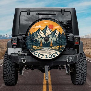 May include: Black Jeep with a spare tire cover featuring a mountain and forest landscape design. The cover has a sunset with the text "GET LOST" in white. The vehicle is on a paved road.